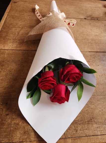Three red roses with foliage, beautifully gift wrapped in white paper with a brown ribbon.